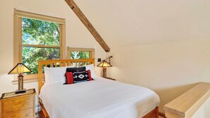 A bedroom with a wooden bed, two bedside lamps, a nightstand, and a window with a view of greenery.