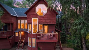 A multi-level wooden cabin with a balcony, large windows, and a view of the surrounding forest.