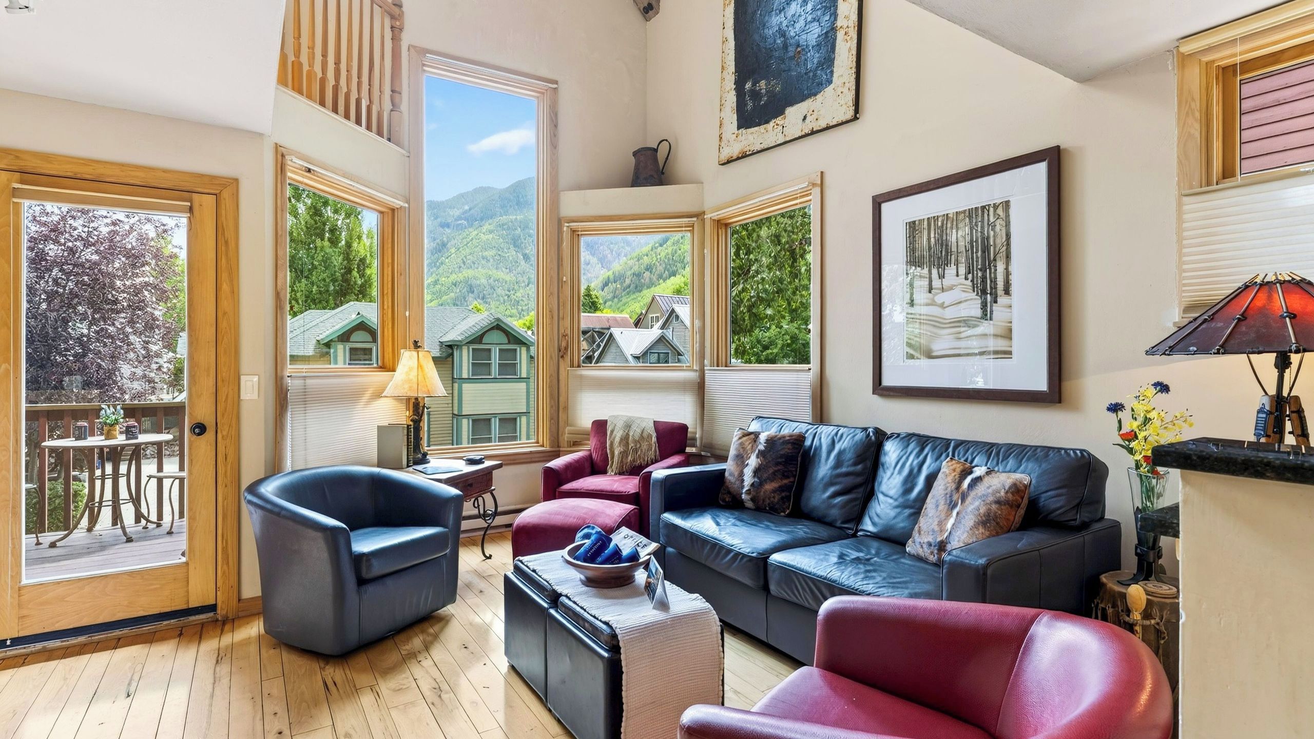 Un salon spacieux avec un grand canapé, un fauteuil rouge et un fauteuil bleu. Deux fenêtres offrent une vue sur une montagne et une maison.