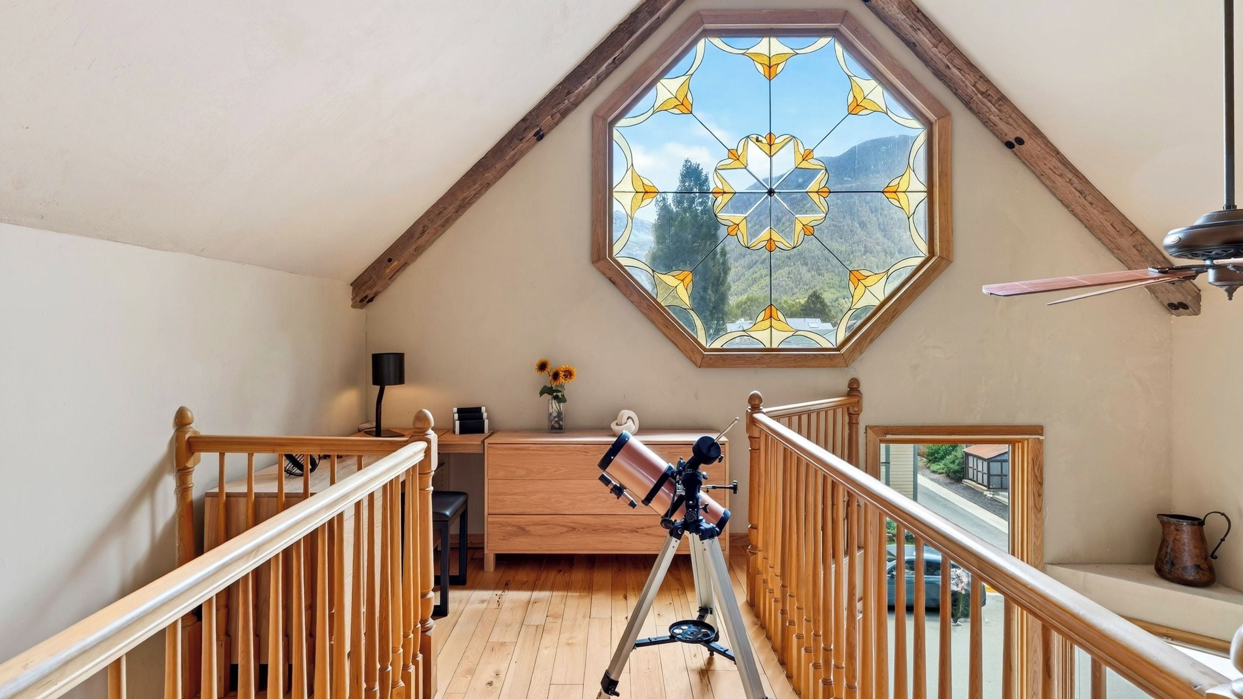 Una habitación con un escritorio de madera, un telescopio y un amplio ventanal octogonal con vistas a la montaña.