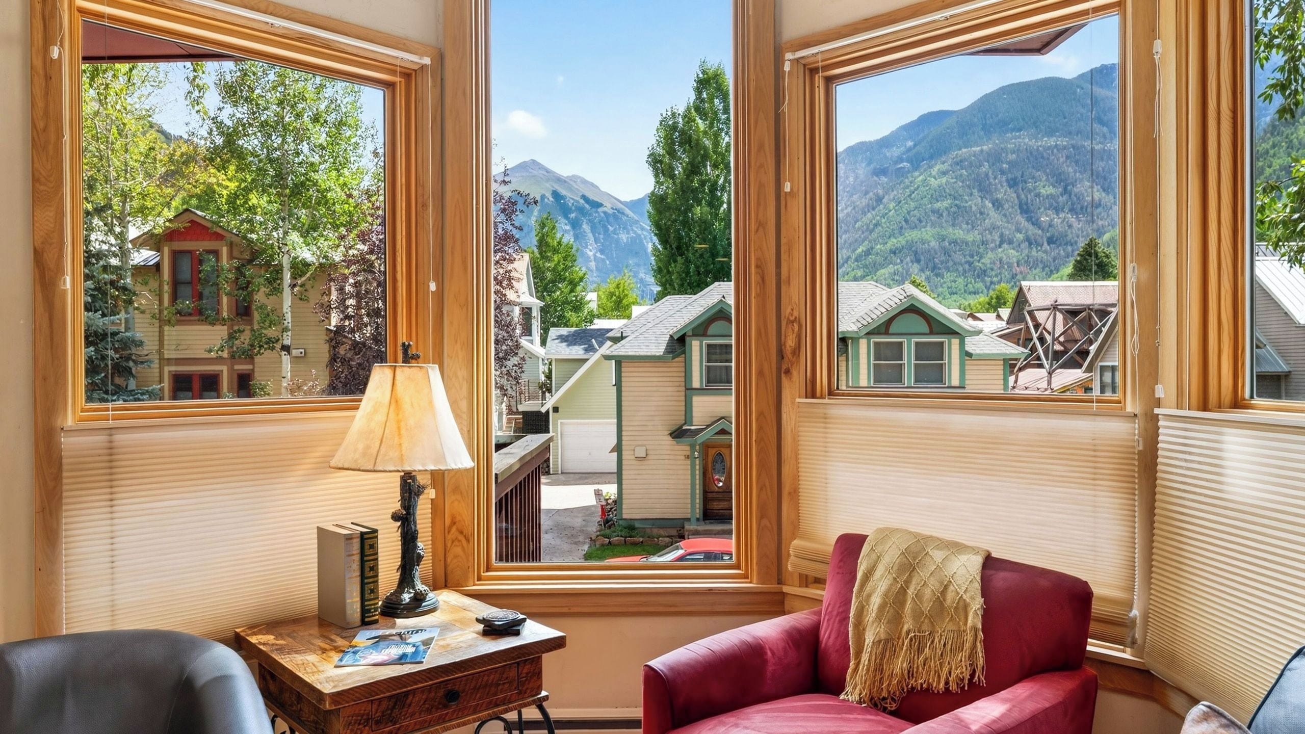 A room with a view of a mountain landscape, a red chair, a wooden table with a lamp, and a book.