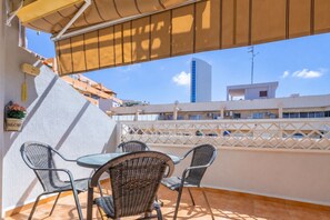 Uma varanda externa com mesa e cadeiras, vista para edifícios e céu claro.