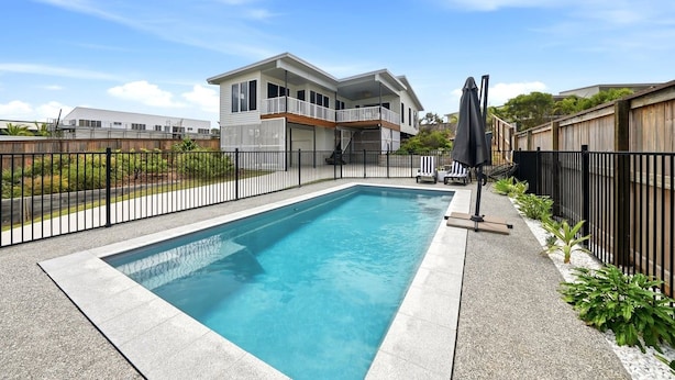 Piscina retangular com cerca de metal preta, área de varanda coberta com móveis de exterior e uma casa de dois andares ao fundo.