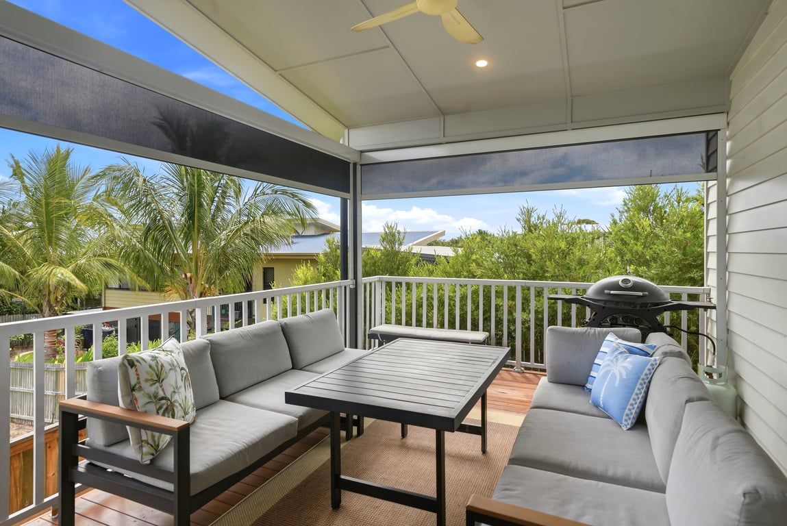 An outdoor patio with a sofa, table, and grill area, surrounded by a white railing and palm trees.