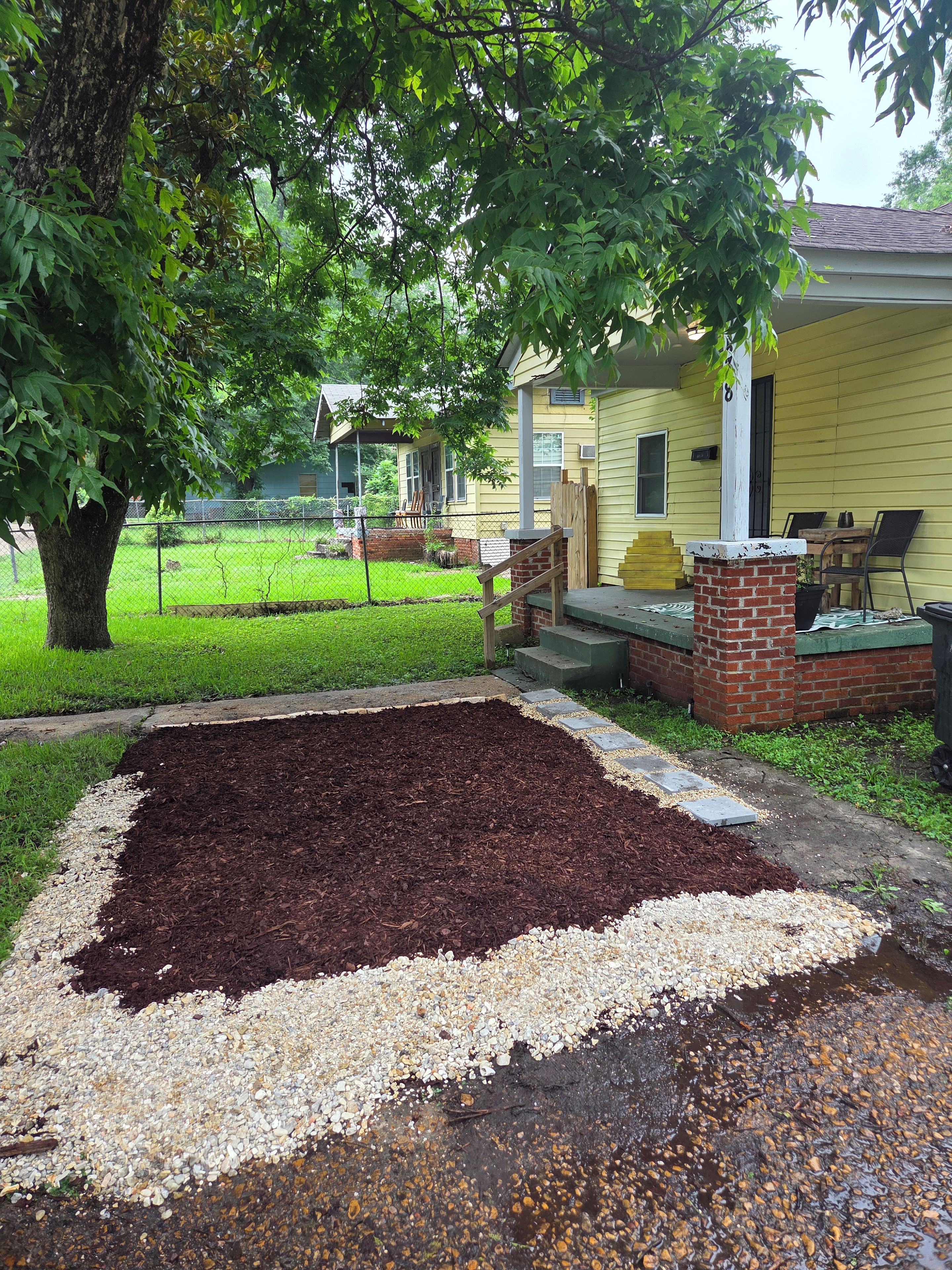 A residential property with a yard, a tree, a brick planter, and a house with a porch.