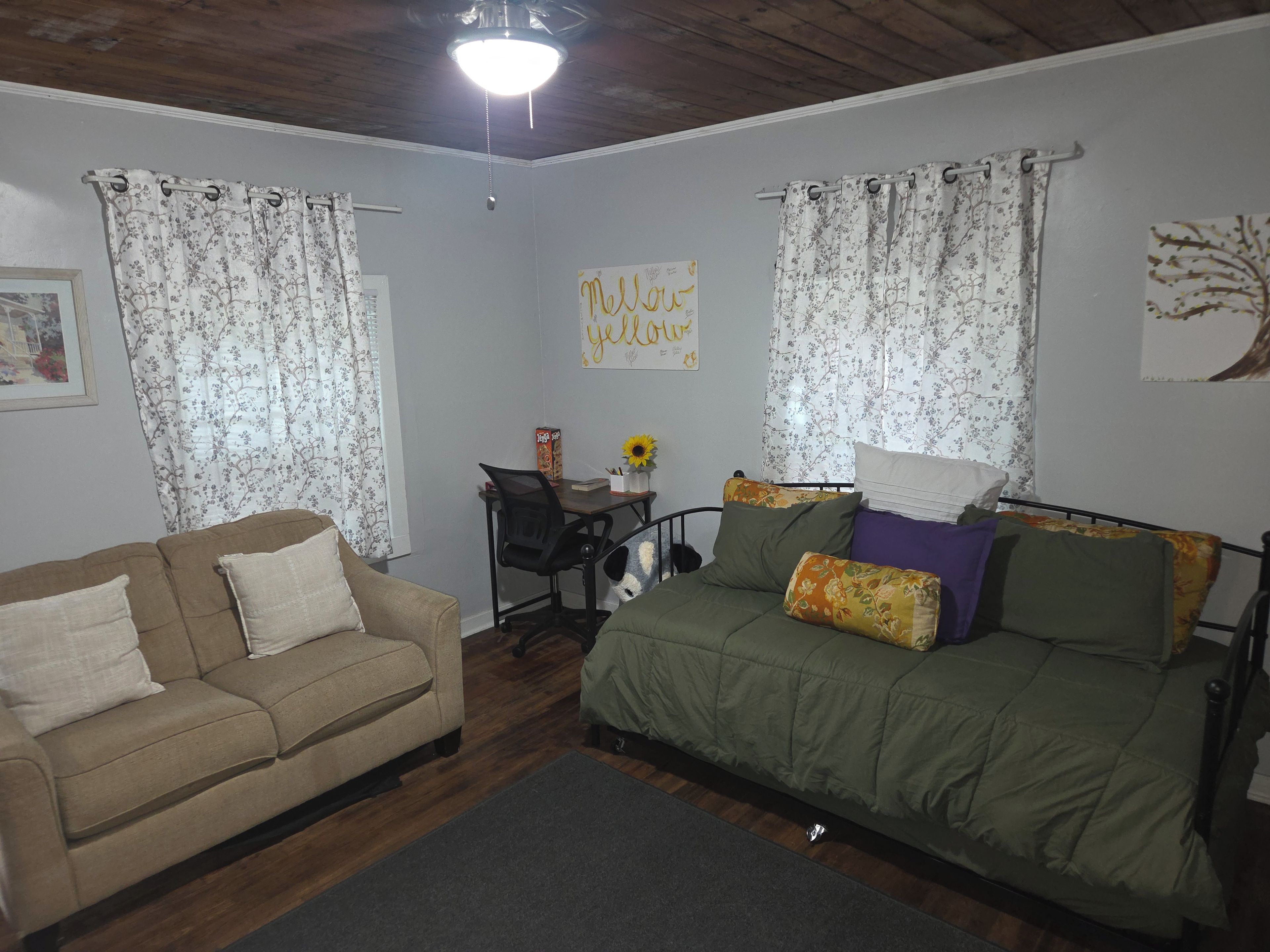 Una habitación con sofá, cama con almohadas florales, escritorio con silla y ventilador de techo.