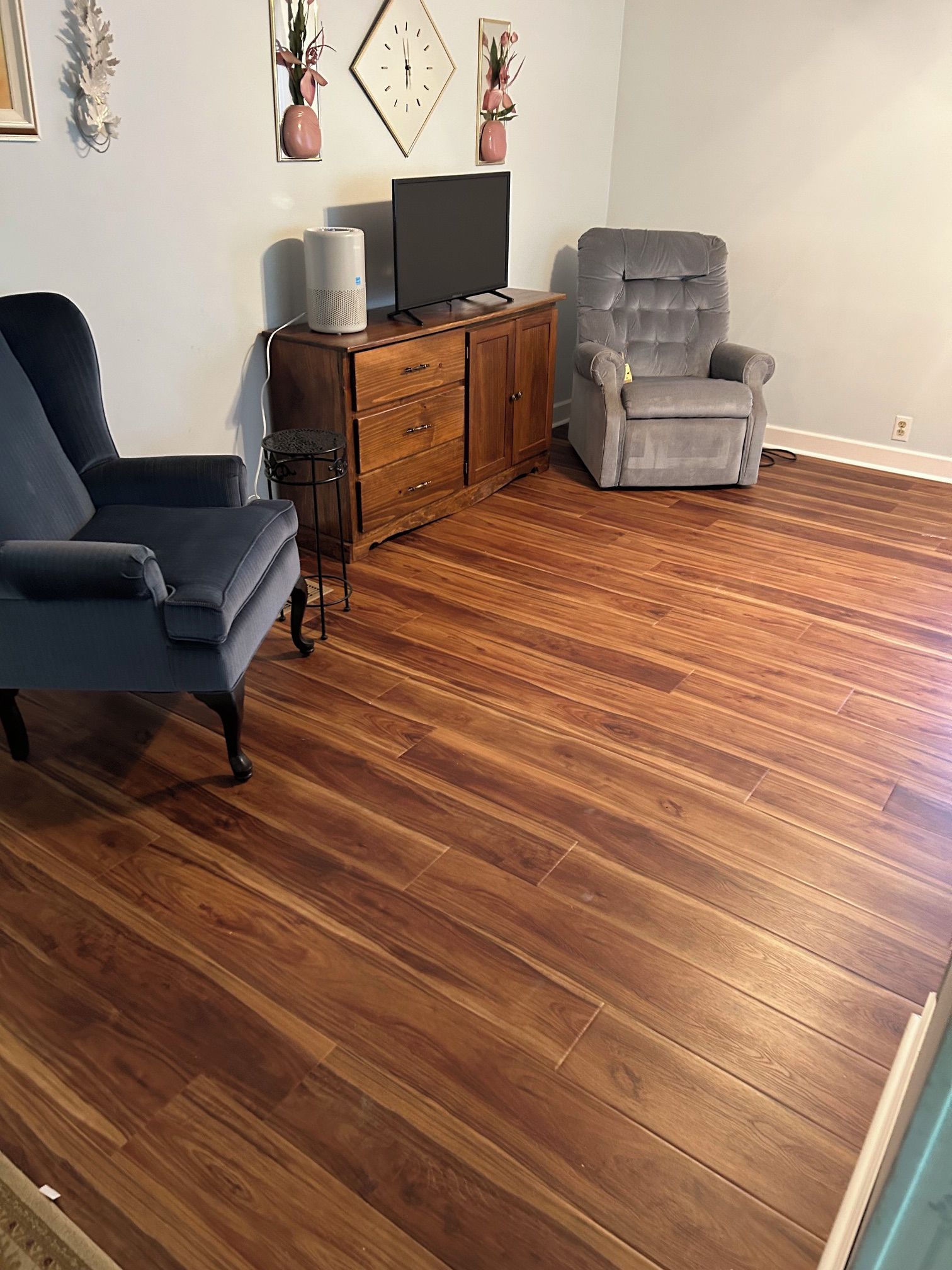 Una sala de estar con un sillón azul oscuro, un sillón reclinable gris, un mueble de madera con un televisor y jarrones decorativos en la pared.
