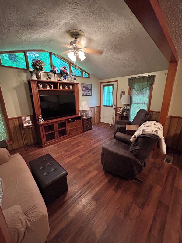A living room with a television, wooden furniture, a ceiling fan, and a window with blinds.
