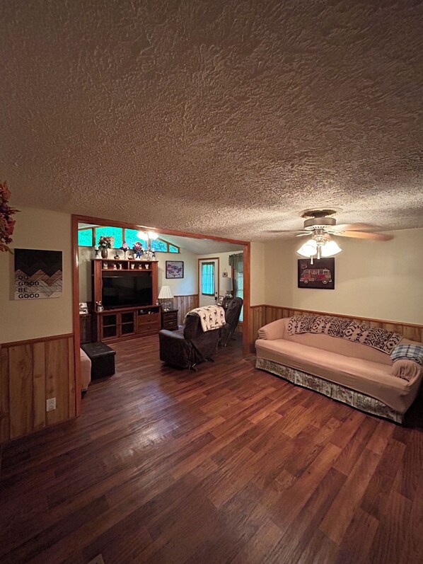 A living room with a wooden floor, a sofa, a television, and a ceiling fan. - Granny's Creekside Lodge 3-bedroom with WiFi & AC in charming Bluefield (Bluefield)