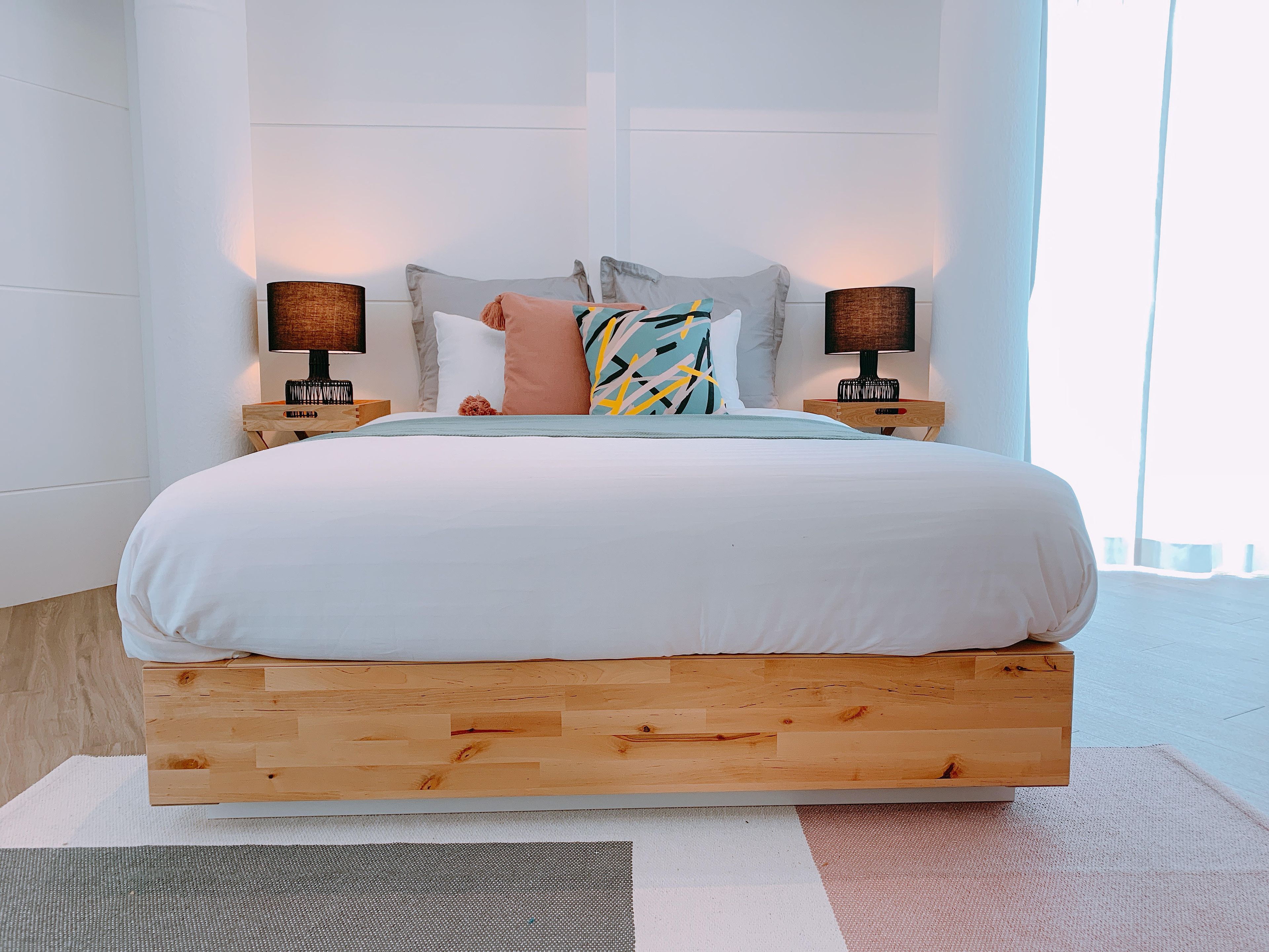 A bed with a wooden frame, white bedding, and a colorful pillow. There are two bedside lamps on wooden nightstands.