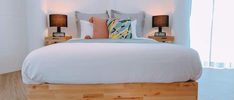 A bed with a wooden frame, white bedding, and a colorful pillow. There are two bedside lamps on wooden nightstands.