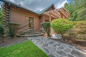 Una baita di legno con un sentiero in pietra che conduce all'ingresso, circondata da vegetazione e un muro di pietra.