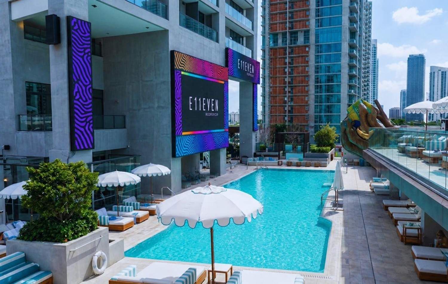 Un espace piscine sur le toit, équipé de chaises longues, de parasols et d’un grand panneau numérique affichant le nom « E11EVEN ROOF ».