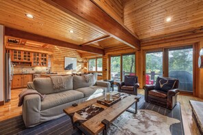 Geräumiges Blockhäuschen-Interieur mit einer großen Holzbalkendecke, einer gemütlichen Sitzecke, einer Küche mit Holdekor-Schränken und großen Fenstern mit Blick ins Freie.