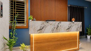 A reception desk with a marble top and wooden base, flanked by potted plants and a window with yellow blinds.
