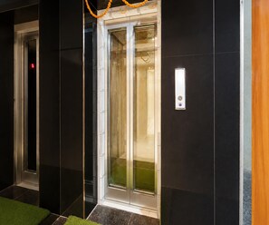A modern elevator with glass doors, a visible emergency exit sign, and a control panel on the wall. - Deluxe Kamala Resort (Bhubaneshwar)