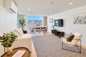 A modern living room with a sofa, a coffee table, a TV, and a dining area with chairs and a table. - Hyde Lane Stay - 3BR with Pool, Gym & Views (Wellington)