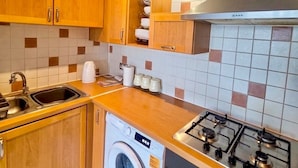 Cocina compacta con armarios de madera, lavadora integrada, estufa a gas y campana extractora de acero inoxidable.
