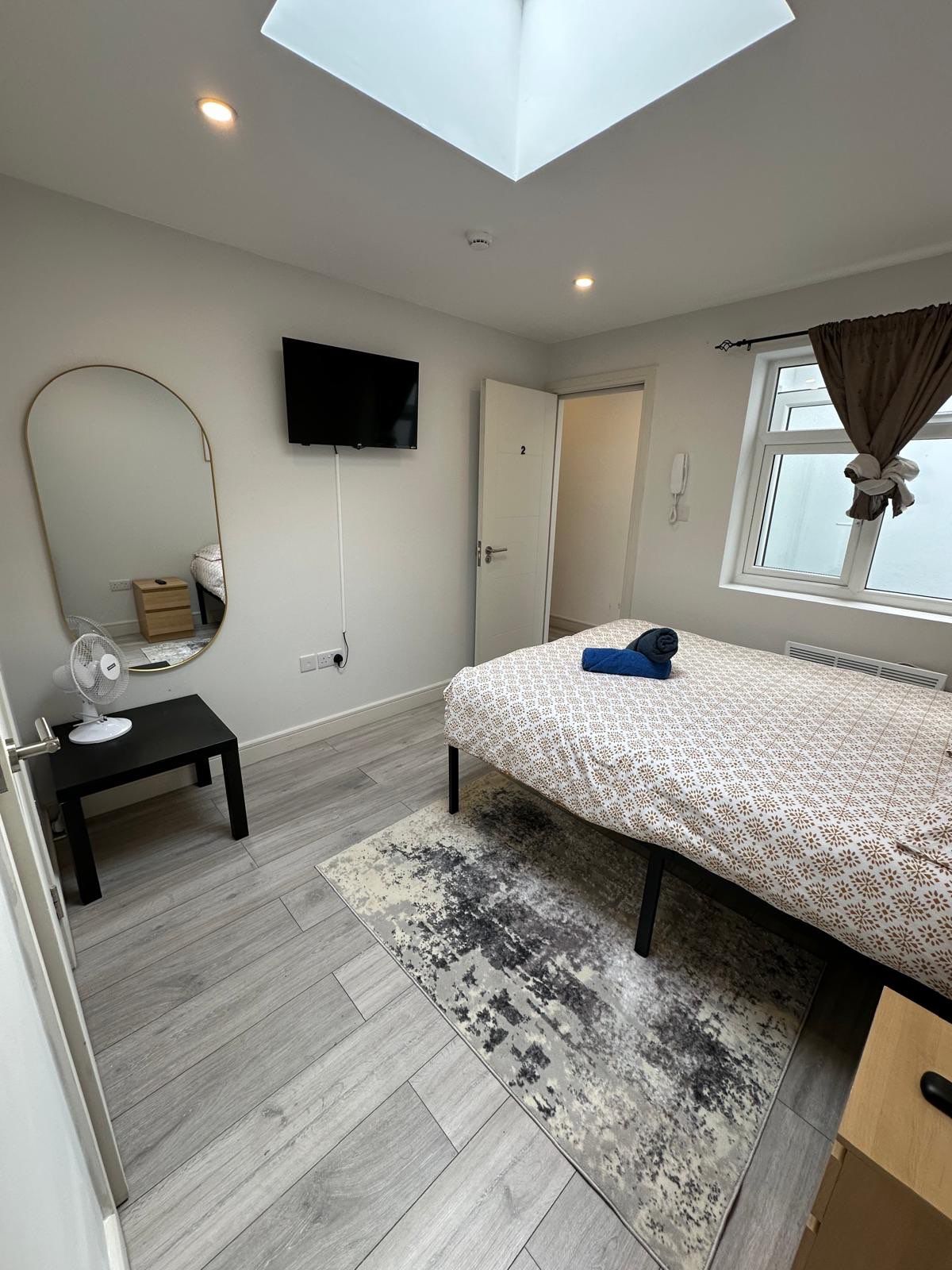 A bedroom with a bed, a mirror, a TV, a small table with a fan, a window with curtains, and a skylight.