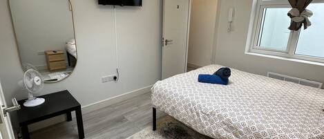 A bedroom with a bed, a mirror, a TV, a small table with a fan, a window with curtains, and a skylight.