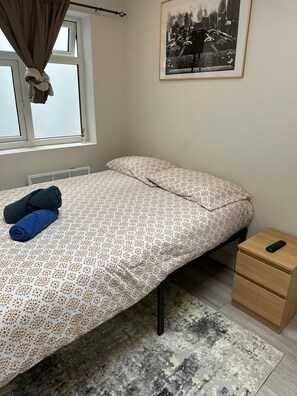 Ein Einzelbett mit einem gemusterten Überwurf, ein Nachttisch mit Fernbedienung, ein Fenster mit Vorhängen und ein gerahmtes Bild an der Wand.