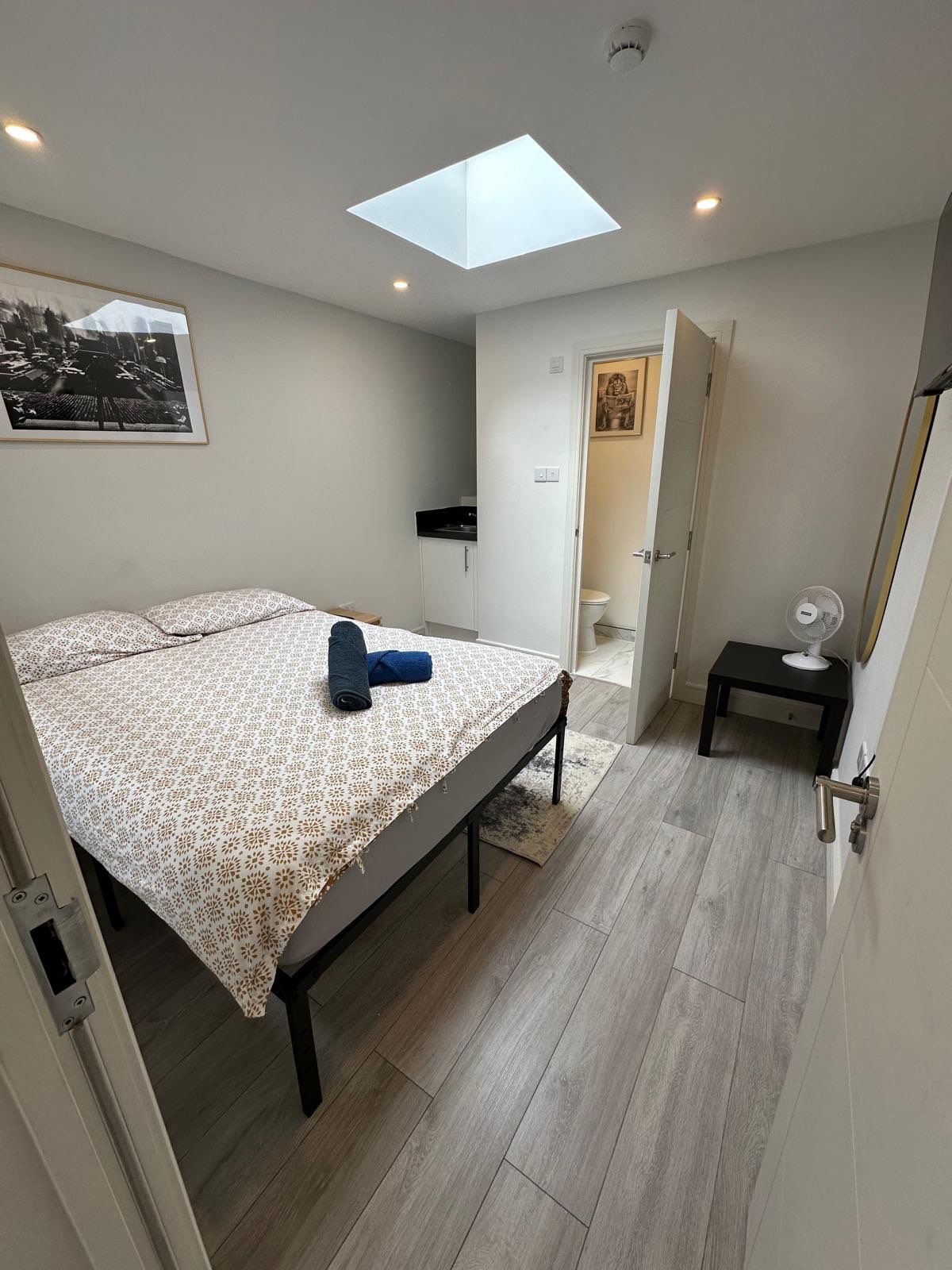 A bedroom with a skylight, a bed with a patterned bedspread, a small bedside table with a fan, and a door leading to a bathroom.
