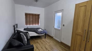 Een kleine, eenvoudig ingerichte kamer met een bed, een houten garderobe, een radiator en een raam met zonwering.
