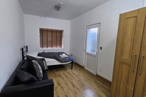 A small, sparsely furnished room with a bed, a wooden wardrobe, a radiator, and a window with blinds.