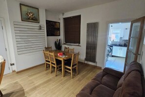 A living room with a brown sofa, a dining area with a wooden table and chairs, a radiator, and a kitchen visible through an open door.