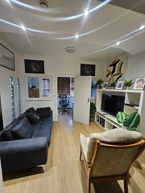 A modern living room with a sofa, a chair, a TV, and a bookshelf. - House Sleeps 10 with Hot Tub and Garden (Romford)