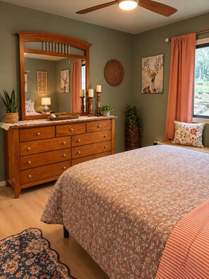 A bedroom with a wooden dresser, a bed with floral bedding, a ceiling fan, and a window with curtains.