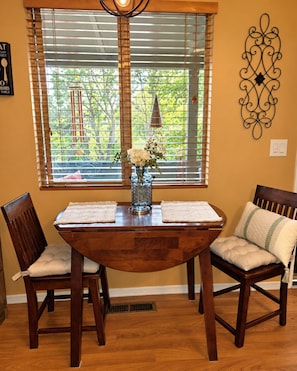 A dining area with a wooden table, two chairs, a vase with flowers, and a window with blinds.