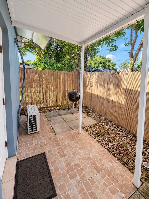 A covered patio area with a barbecue grill, air conditioning unit, and a wooden fence.