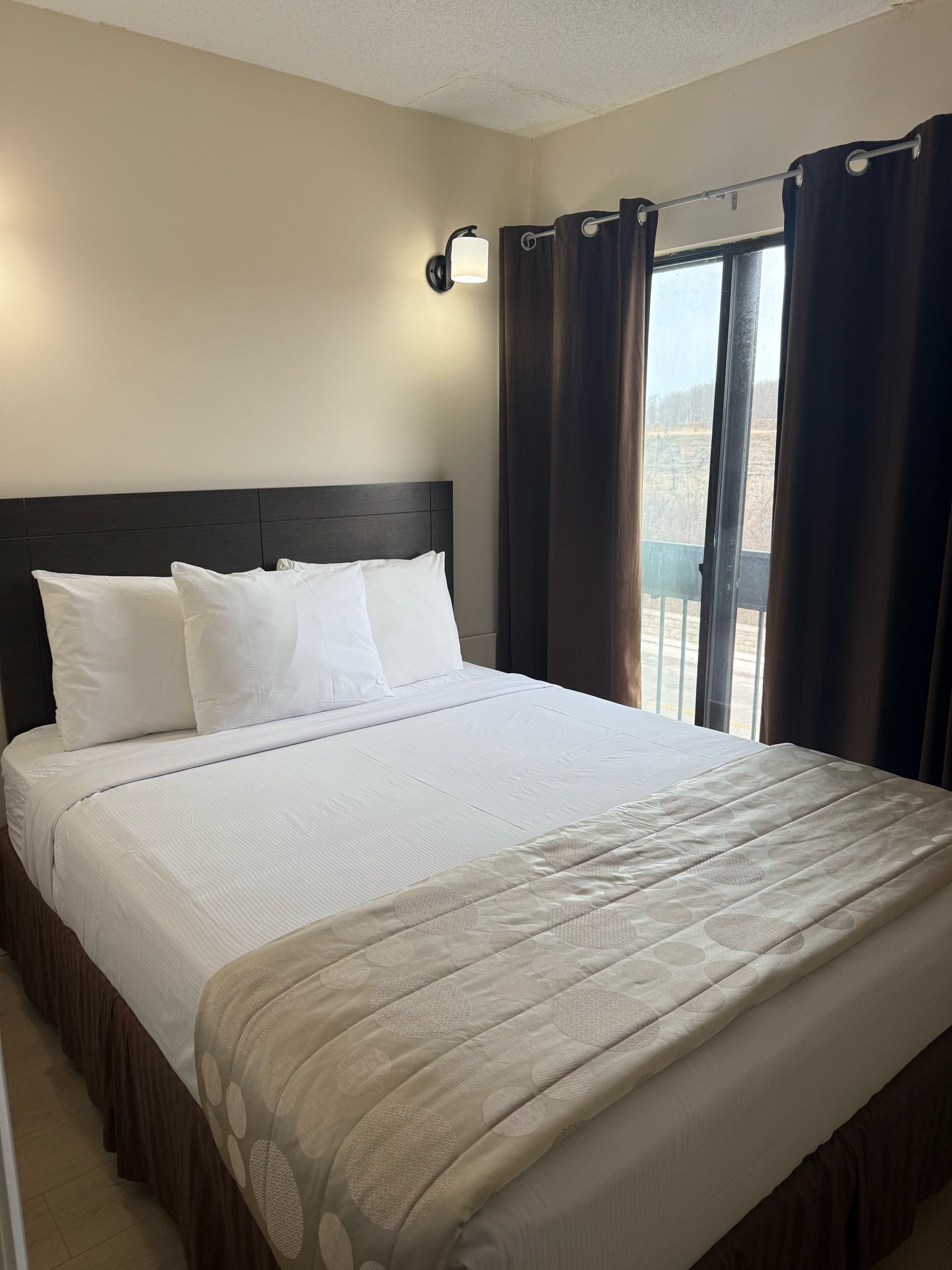 A neatly made bed with white linens and a beige patterned throw, a dark headboard, and a window with curtains.