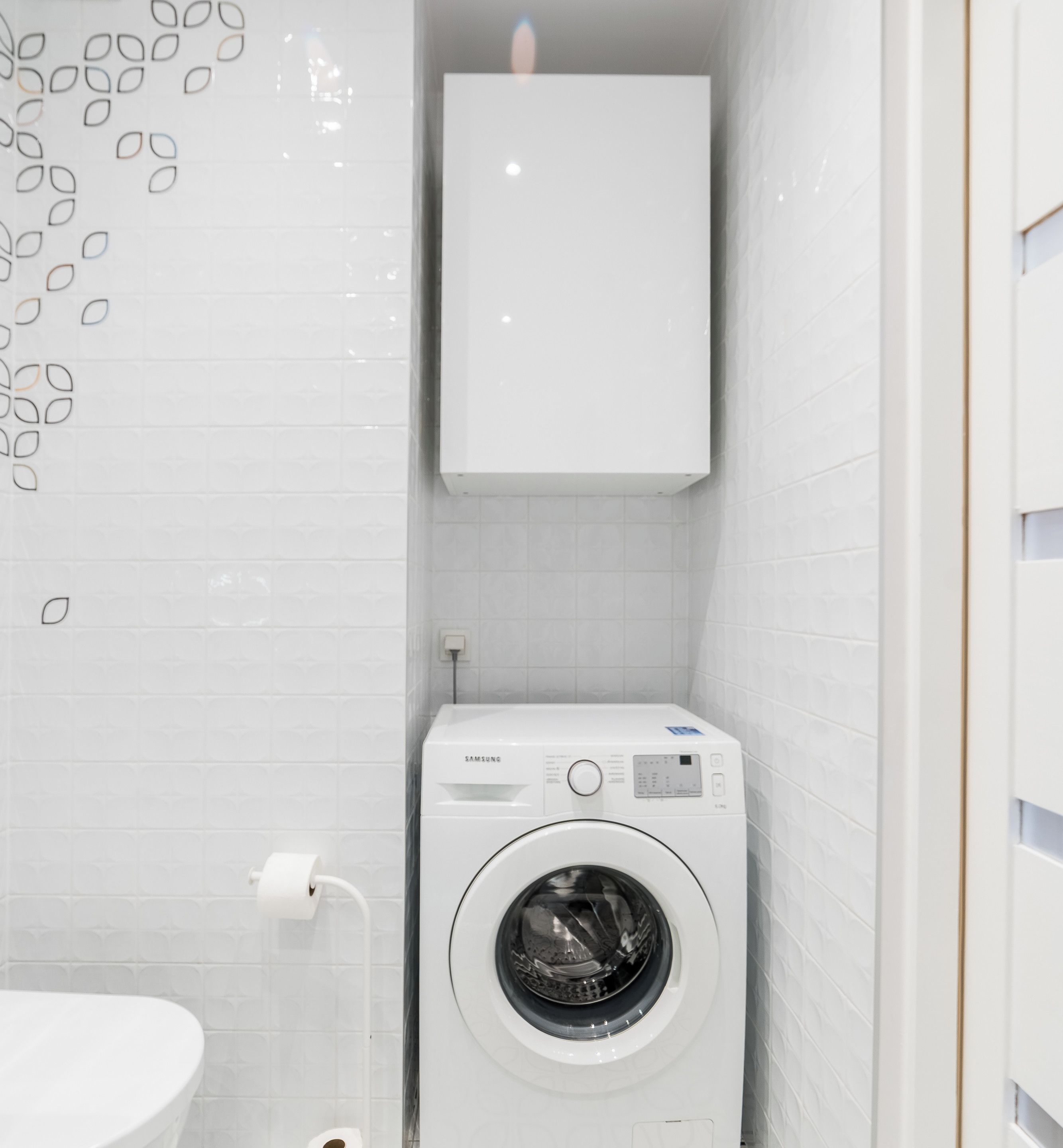 A compact, white laundry room with a front-loading washing machine, a toilet, and a wall-mounted cabinet.
