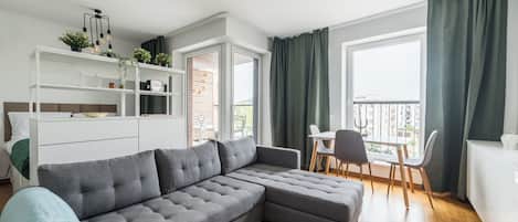 A modern living room with a grey sectional sofa, a white cabinet, a dining table with chairs, and a balcony with green curtains.