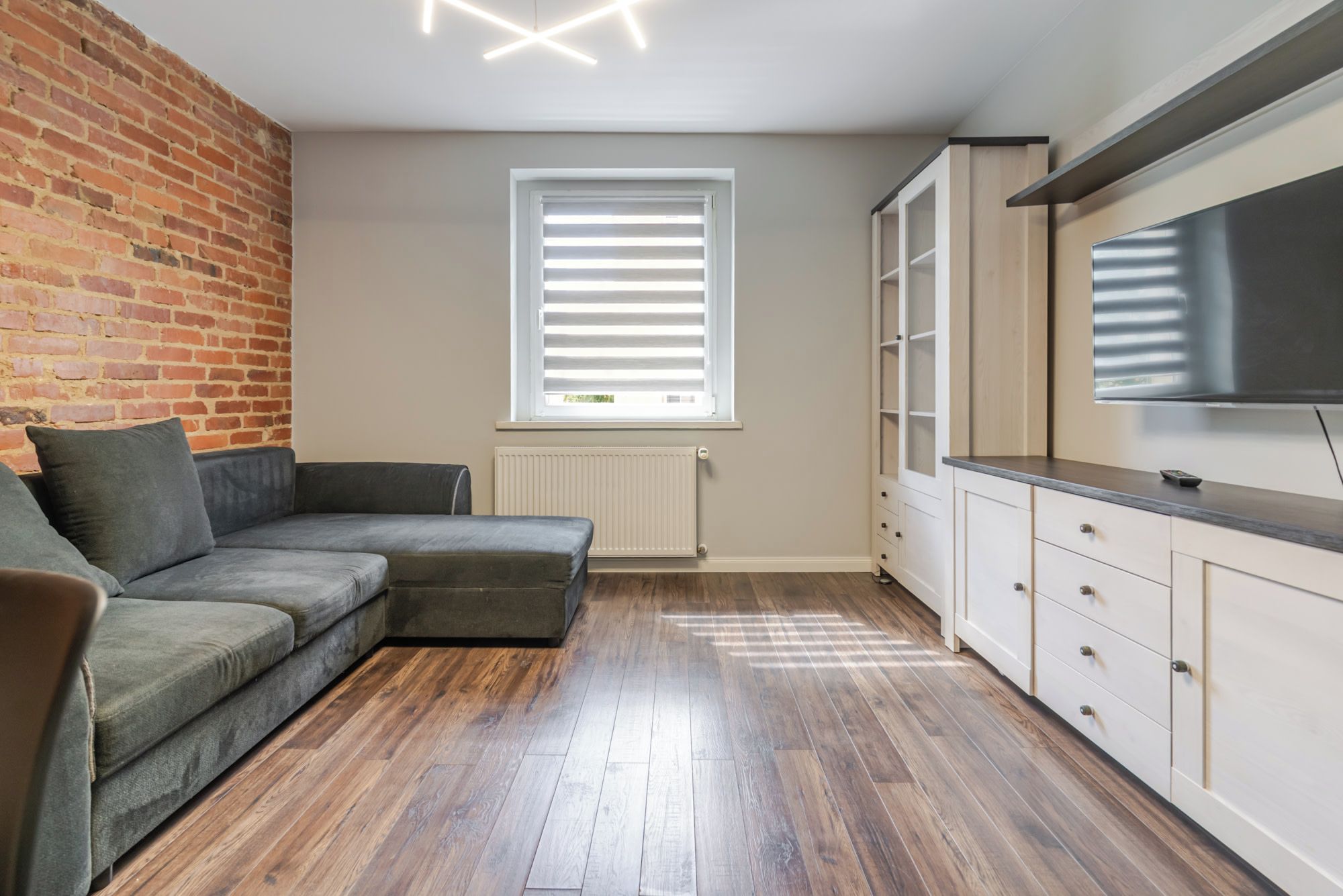 Un salon moderne avec un canapé d’angle, un mur en briques, une fenêtre avec des stores, un radiateur et un meuble télé intégré.