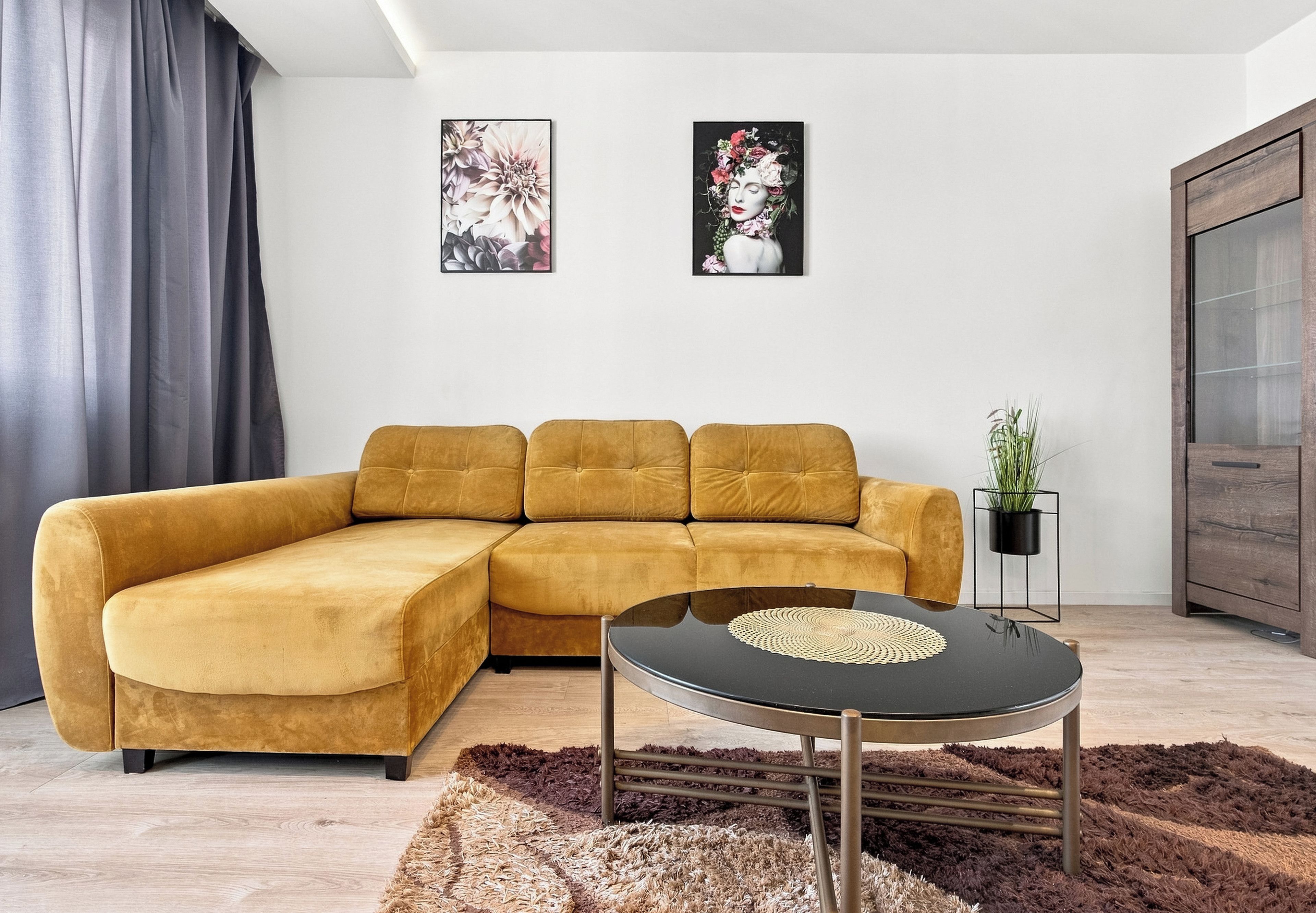 Una sala de estar moderna con un sofá seccional color mostaza, una mesa de centro redonda negra y dos cuadros con motivos florales en la pared.