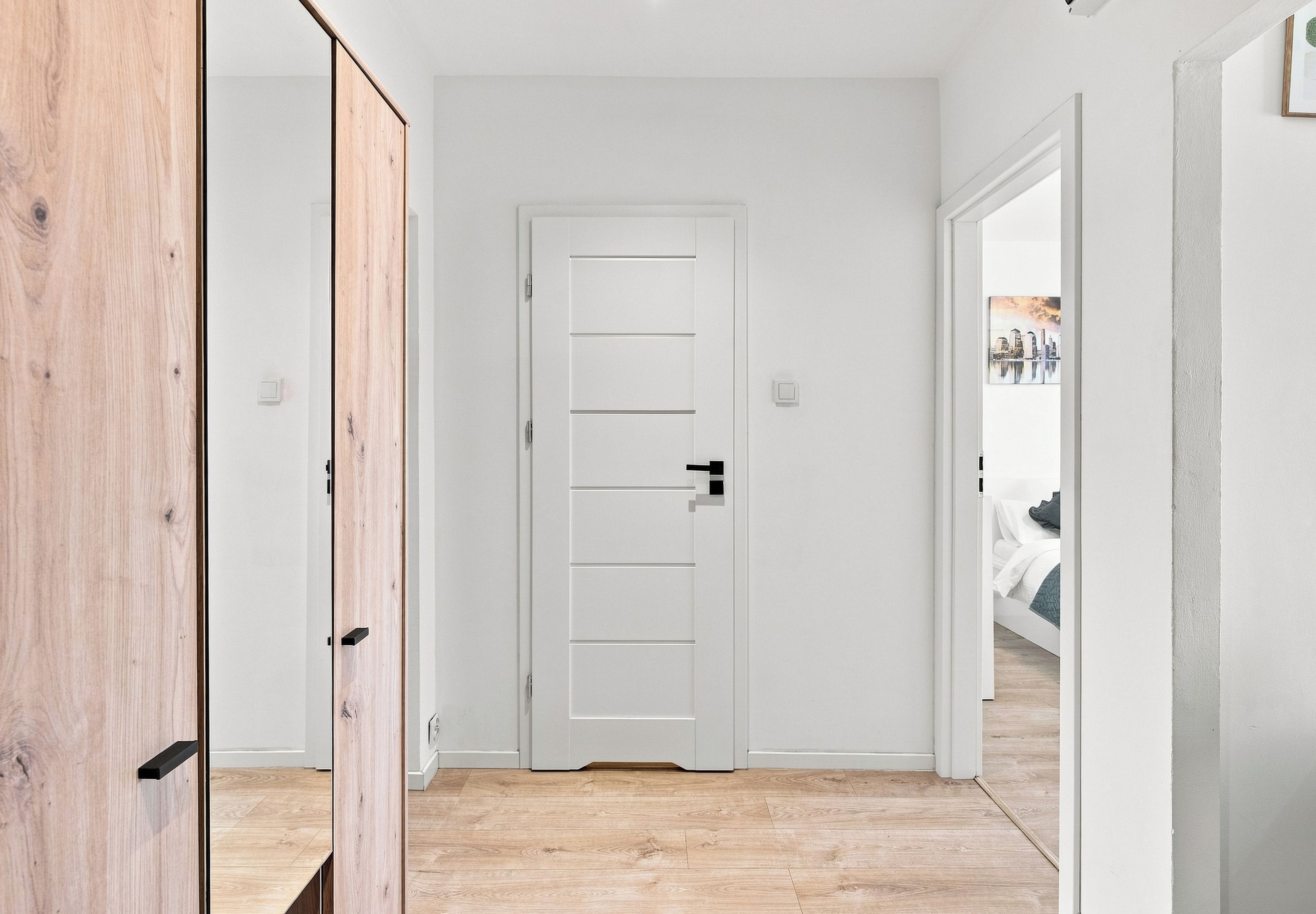 Un pasillo con puerta blanca, suelo de madera y vista de una cama en la habitación contigua.
