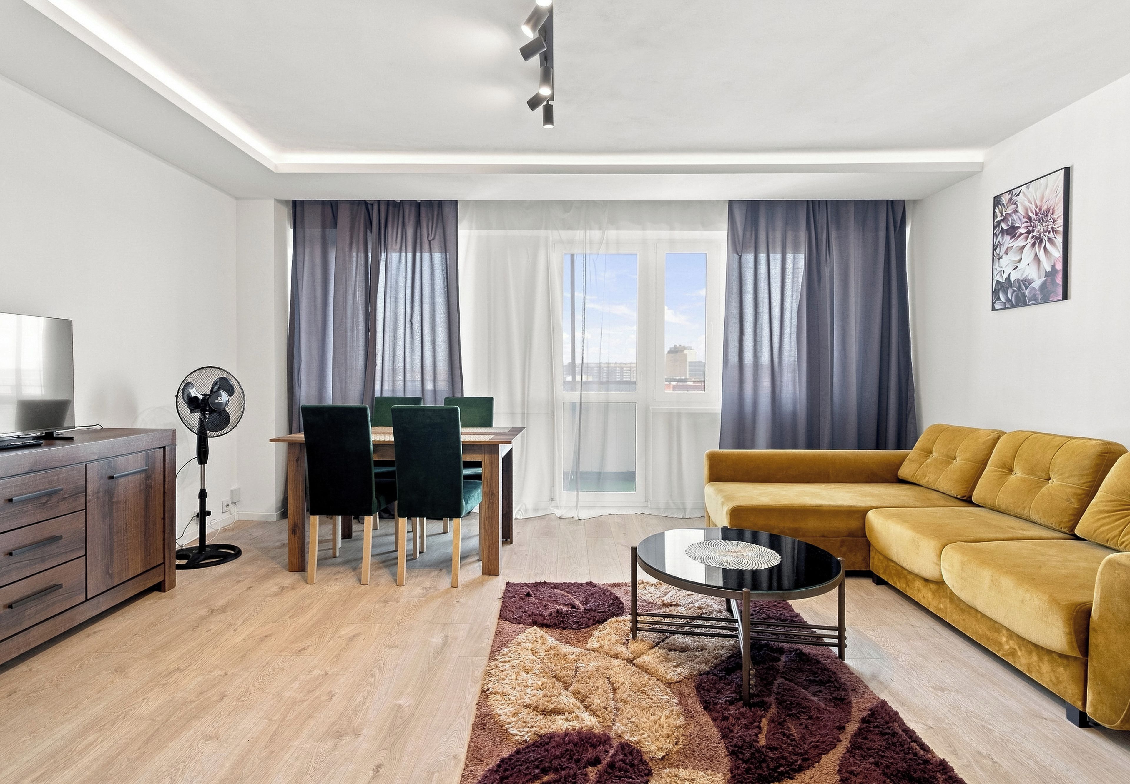 A modern living room with a sofa, a dining table with chairs, a fan, and a large window with curtains.