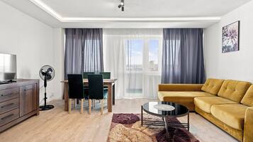A modern living room with a sofa, a dining table with chairs, a fan, and a large window with curtains.