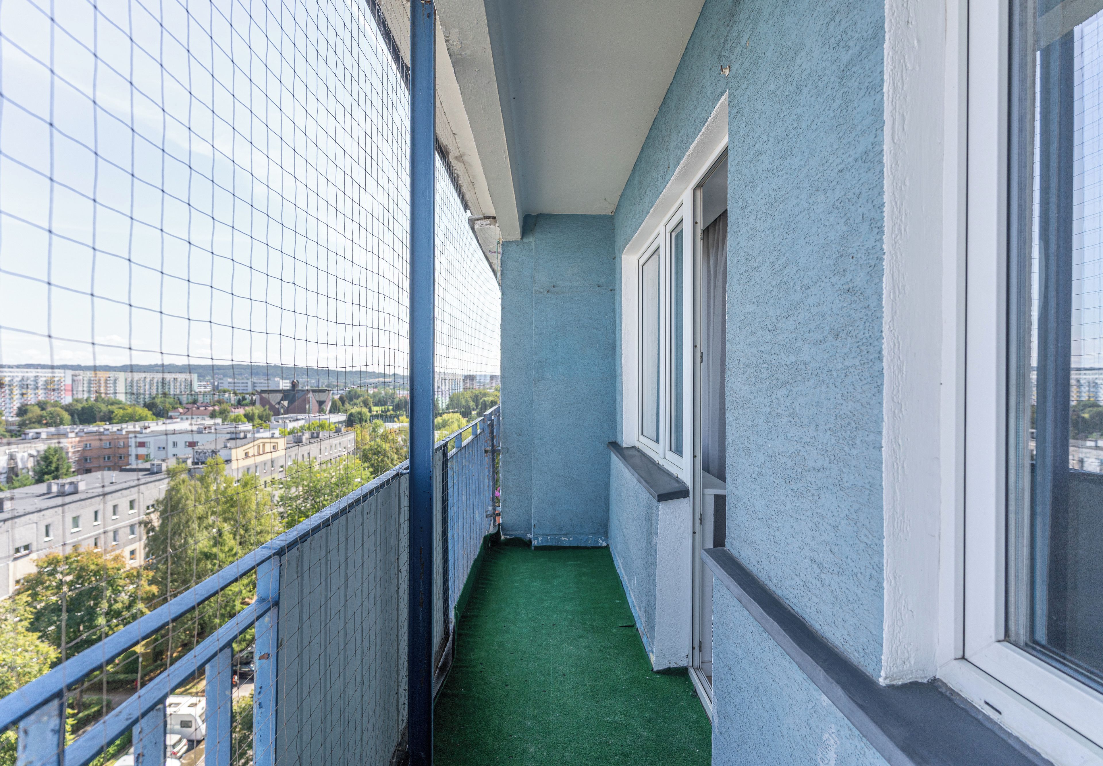 Un balcó estret amb un paviment de gespa artificial verda, barana de metall i vista d'un paisatge urbà amb edificis i arbres.