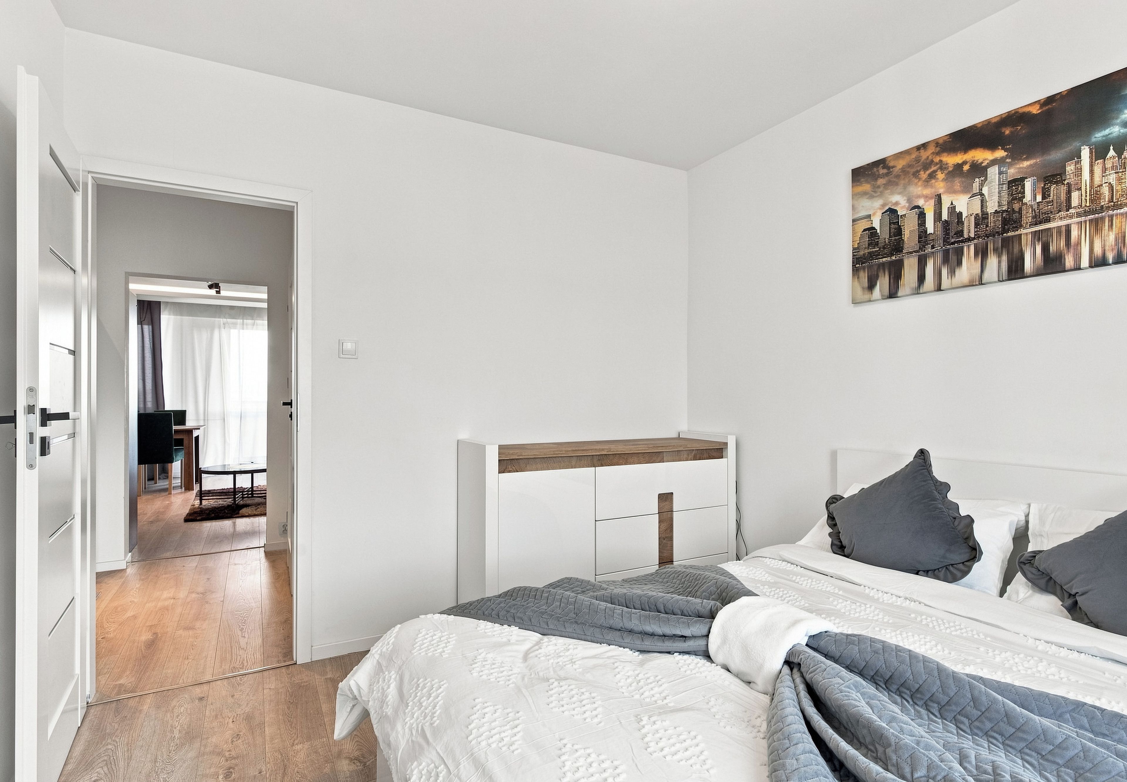 A bedroom with a bed, a nightstand, a painting of a cityscape, and a view of a dining area through an open door.