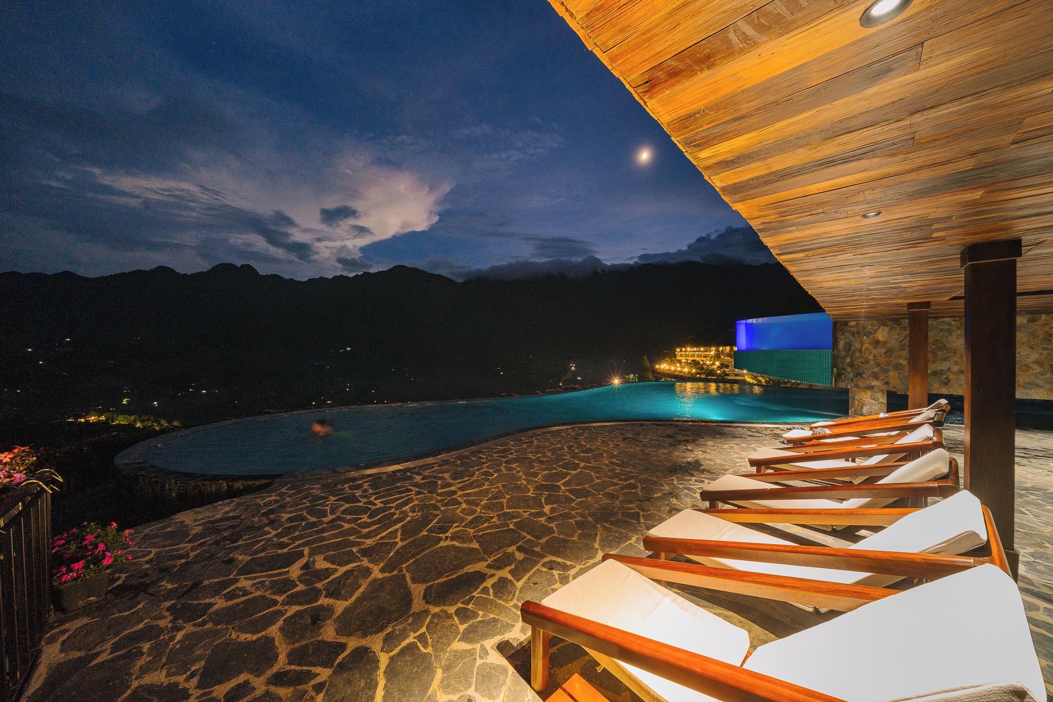 Vista nocturna de un área de piscina con sillones de descanso y terraza de piedra.