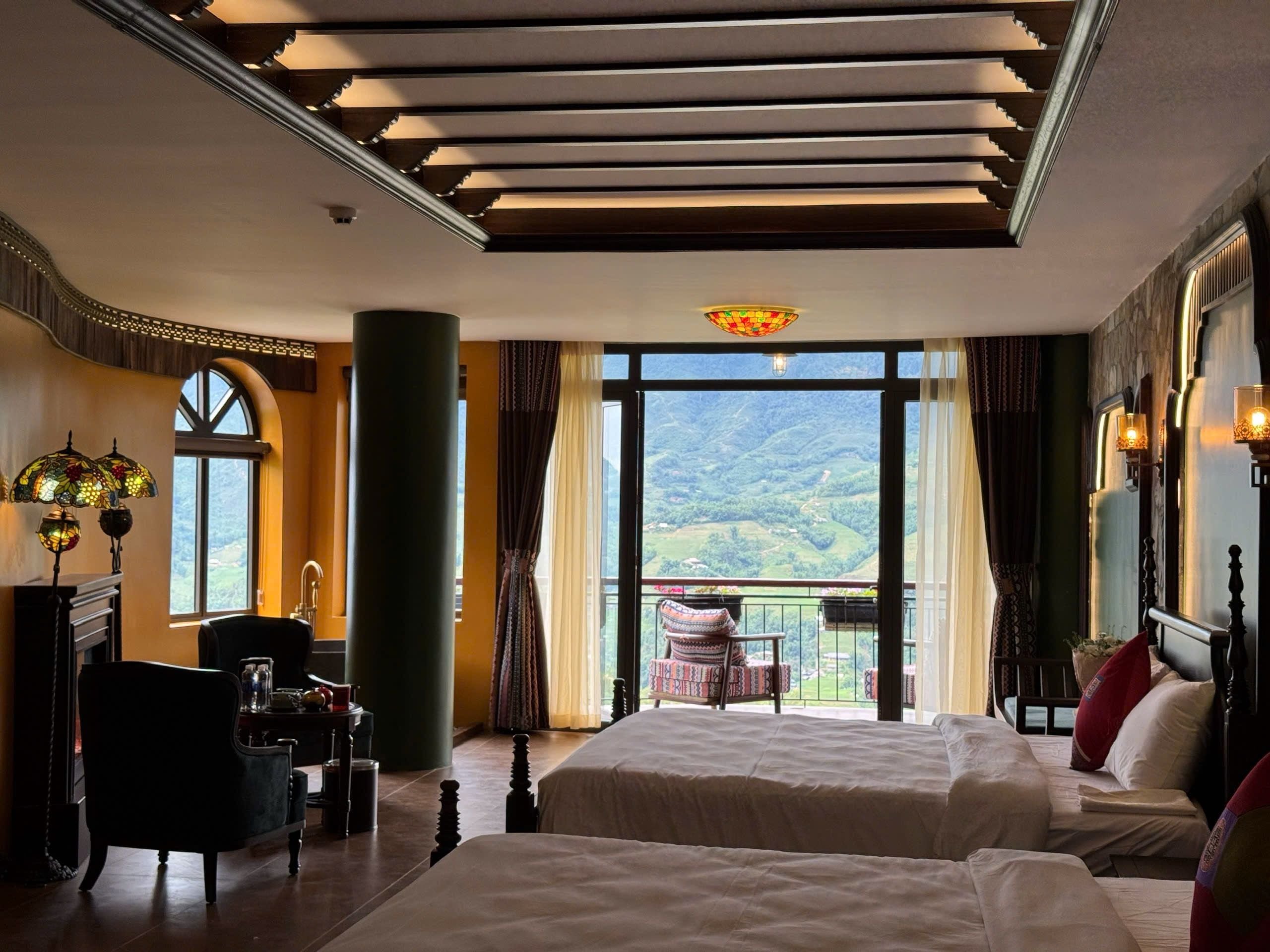 Una camera d'albergo con due letti, un'area salotto con una poltrona, un'ampia finestra vista sulle montagne e un lampadario.