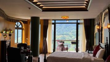 Una camera d'albergo con due letti, un'area salotto con una poltrona, un'ampia finestra vista sulle montagne e un lampadario.