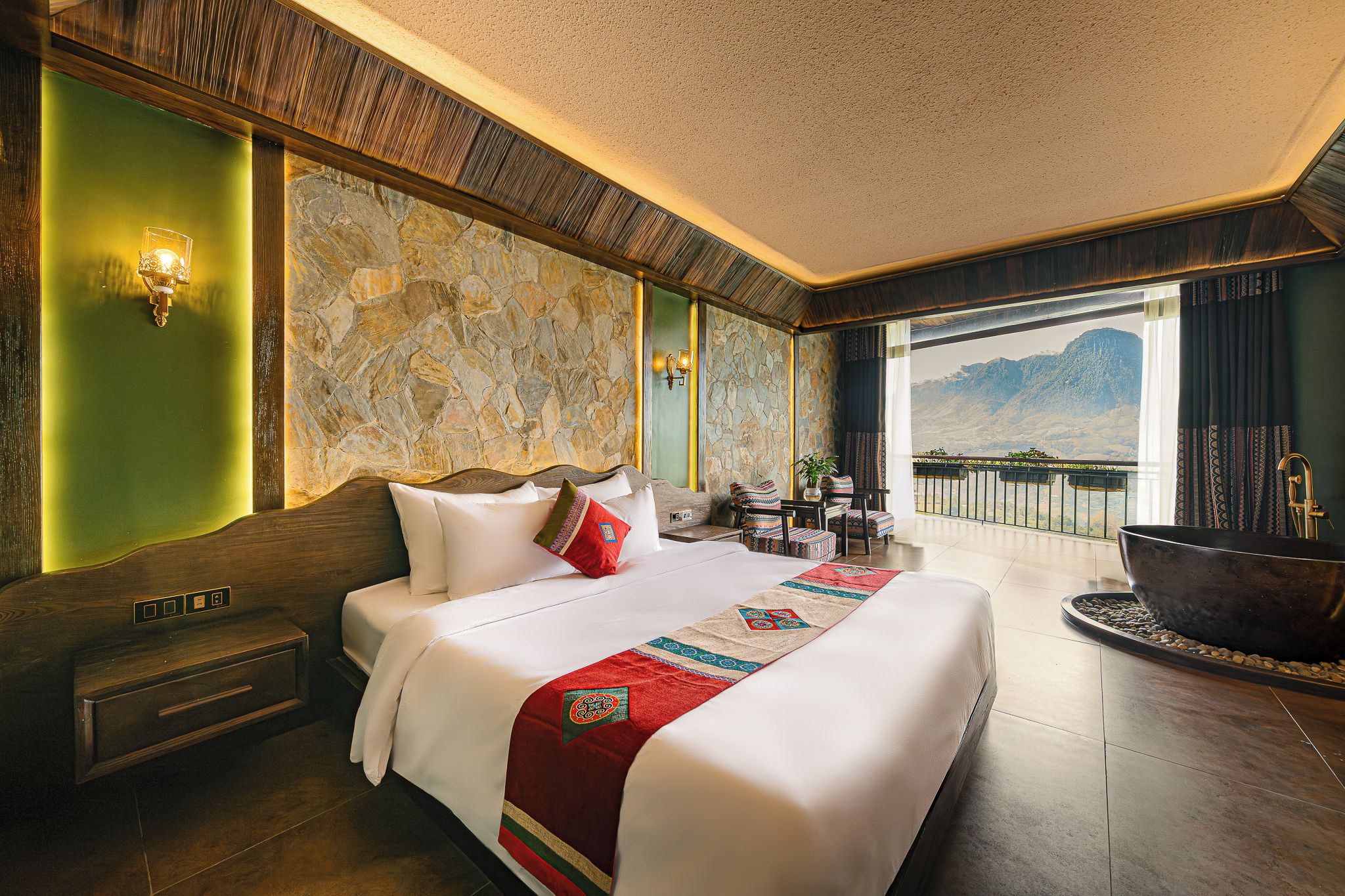 A hotel room with a large bed, a stone wall, a view of mountains, a balcony with a hot tub, and a wooden floor.