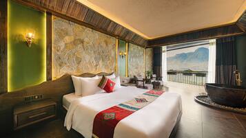 A hotel room with a large bed, a stone wall, a view of mountains, a balcony with a hot tub, and a wooden floor.