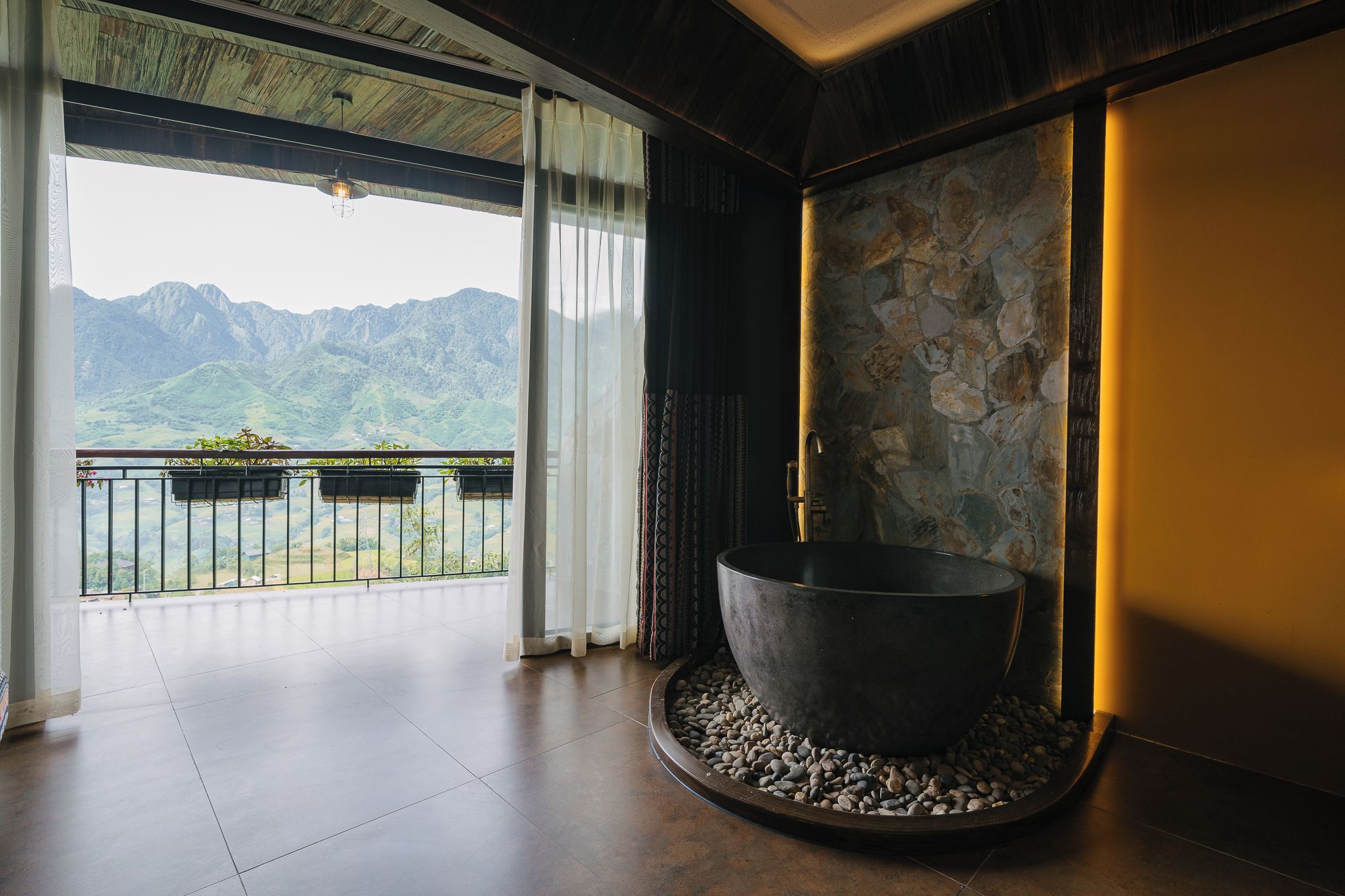 A room with a large stone basin, a balcony with plants, and a view of mountains.