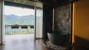 A room with a large stone basin, a balcony with plants, and a view of mountains.